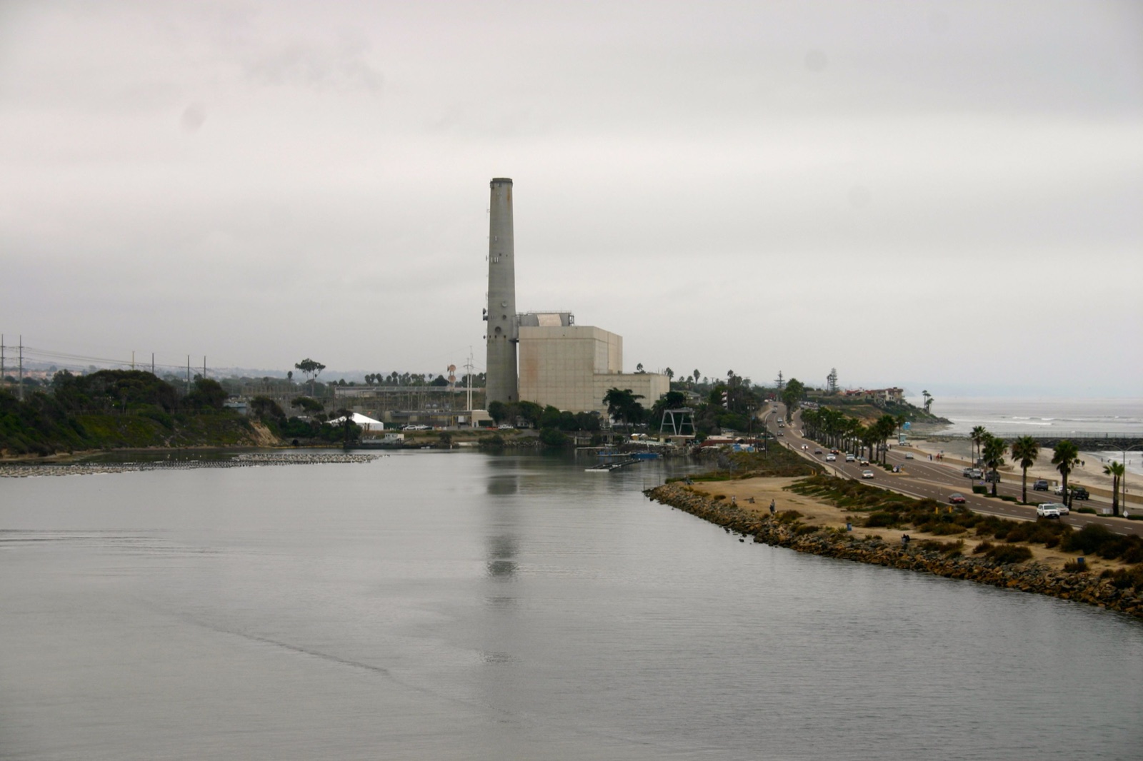 The Encina Power Station photographed across Agua Hedionda Lagoon from Cannon Road, Carlsbad — the shoreline view showing the plant, the intake channel, and the water body that both feeds the desalination plant next door and defines the Coastal Zone constraints on redevelopment.